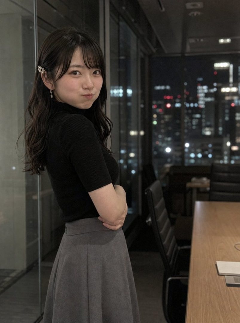 A young woman with long black hair and bangs, wearing a black top and grey skirt, poses in an office at night with a city view in the background.