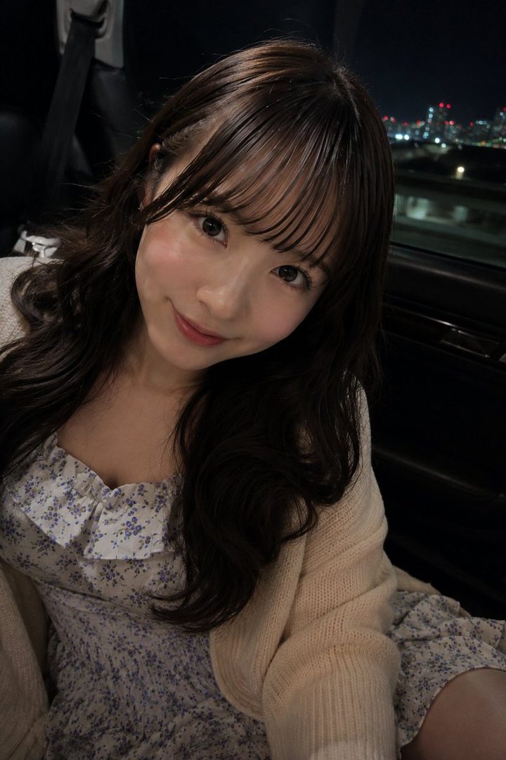 A photorealistic portrait of a young woman with long brown hair and bangs, wearing a floral dress and a cardigan, smiling softly in a car at night with city lights in the background.