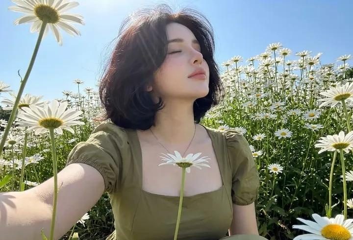 A woman with short dark hair and an olive green dress sits in a field of white daisies under a clear blue sky.