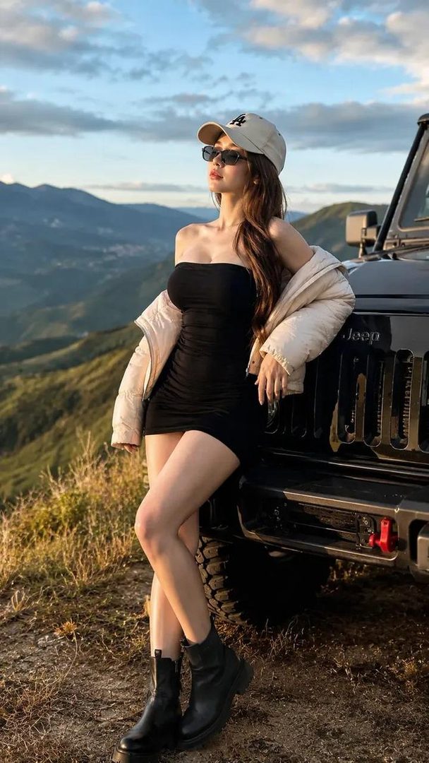A woman in a black mini dress and puffer jacket leans against a Jeep on a mountain road during golden hour.