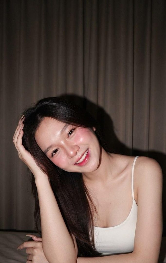 A young woman with long dark hair and a white camisole smiles brightly, with a hand resting on her head, against a brown curtain background.