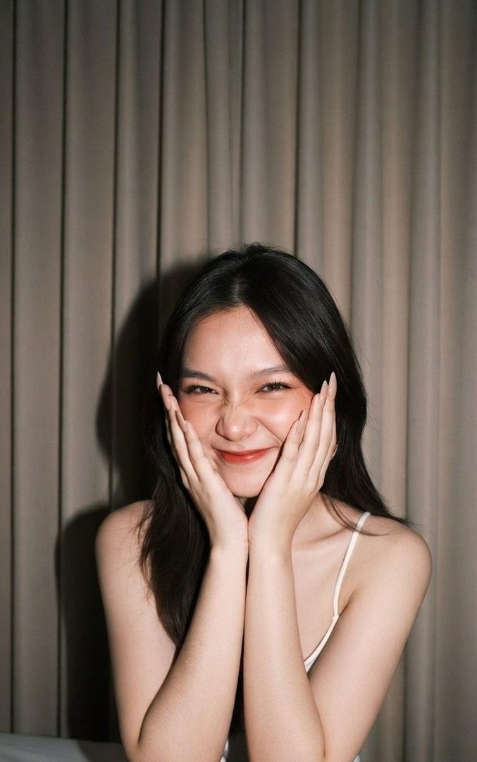 A young woman with long dark hair smiles playfully at the camera, holding her hands to her cheeks against a curtain background.