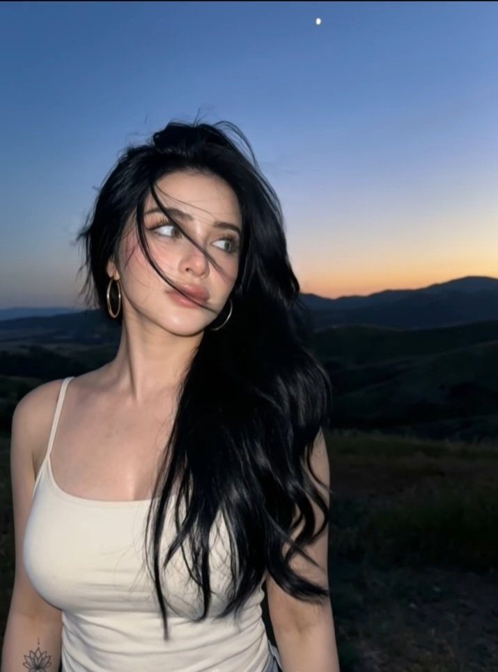 A woman with long black hair and a white tank top looks away into the distance during golden hour in a hilly landscape.