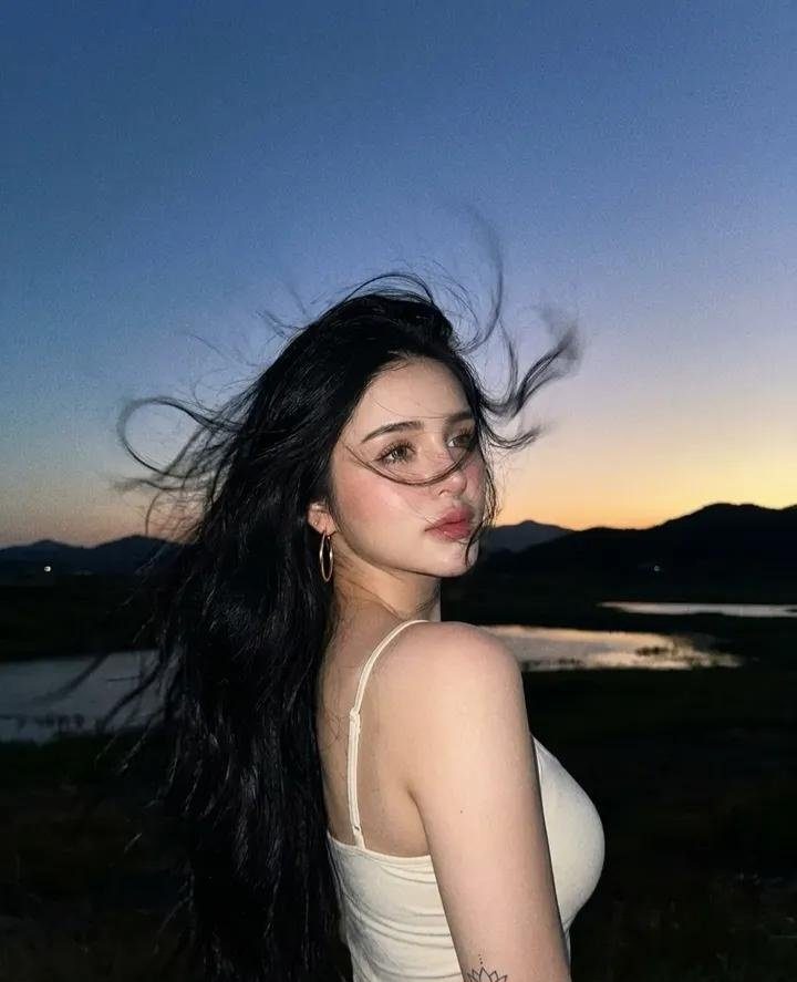 A woman with long black hair and a white tank top looks over her shoulder at dusk with a lake and mountains in the background.