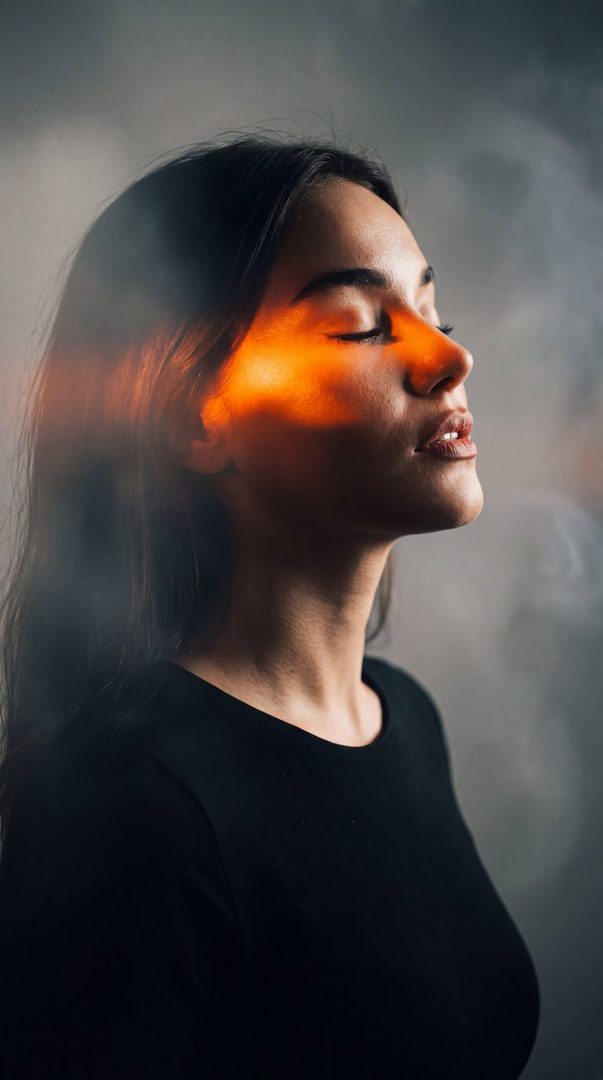A photorealistic portrait of a woman with closed eyes and an orange light effect on her face, set against a smoky gray background.