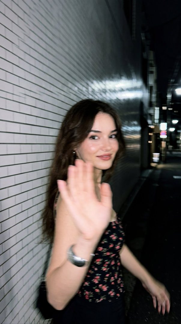A young woman with long brown hair, wearing a floral top, waves at the camera on a dark city street at night.