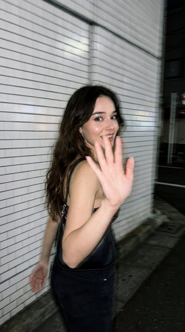A young woman with long brown hair smiles shyly and raises her hand to the camera on a dark city street at night.