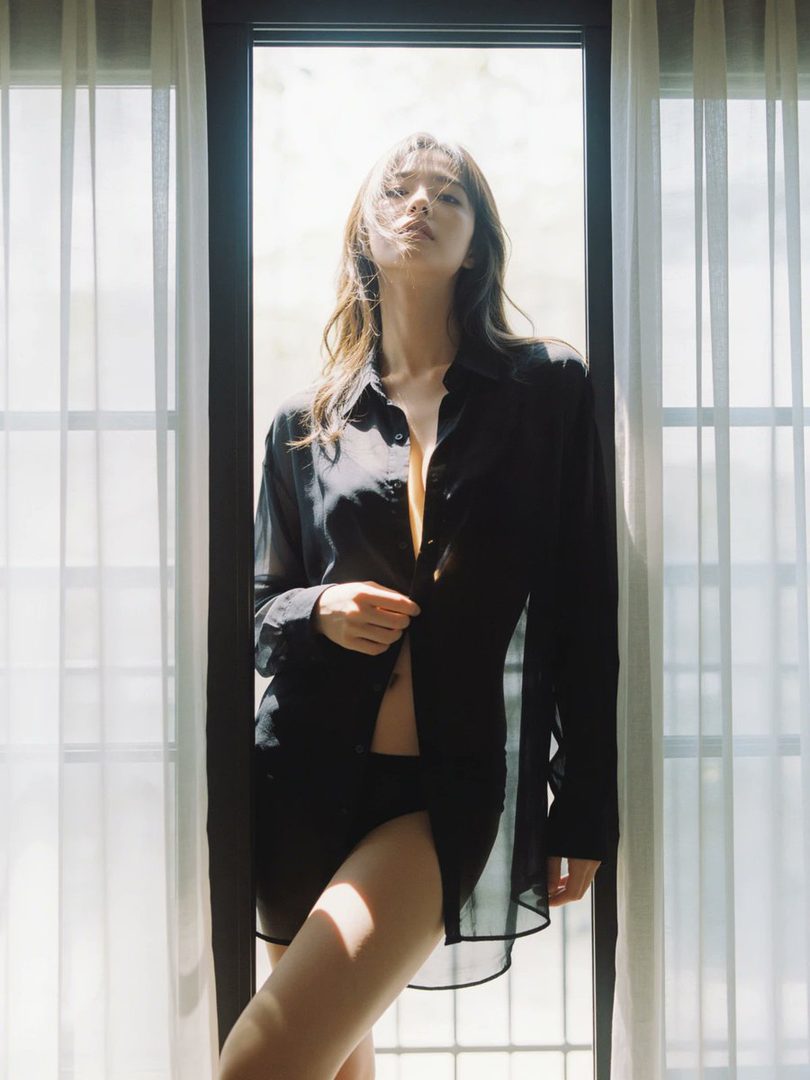 A woman with long brown hair stands in front of a window, wearing an unbuttoned black sheer blouse and black underwear, with light streaming in from behind.