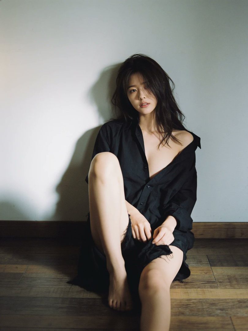 A woman with long black hair sits on a wooden floor, leaning against a white wall, wearing an unbuttoned black shirt.
