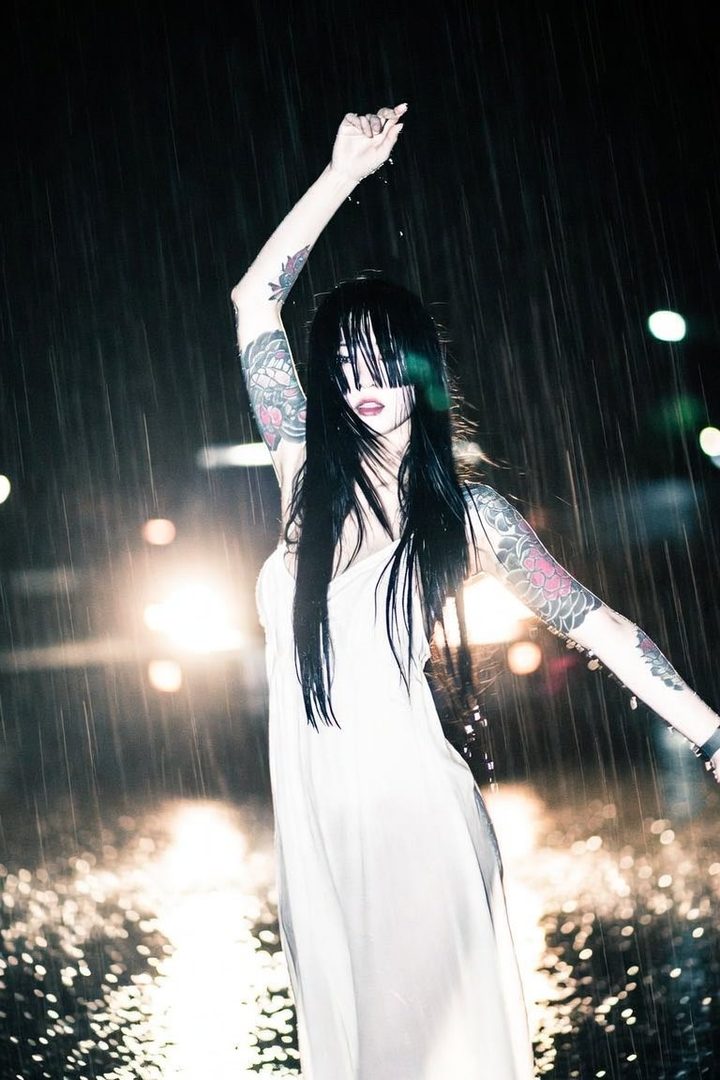 A woman with long black hair and bangs covering her eyes, wearing a white dress, stands in the rain at night with her arm raised.