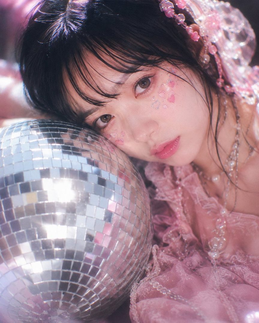 A dreamy portrait of a young woman with black hair and bangs, adorned with pink glitter on her face, resting her head on a disco ball.