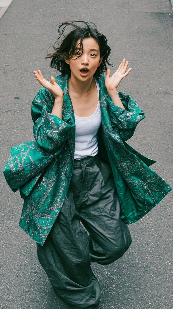 A young Japanese woman with short black hair, wearing a green patterned jacket and dark pants, looks up at the camera with a surprised expression on a street.