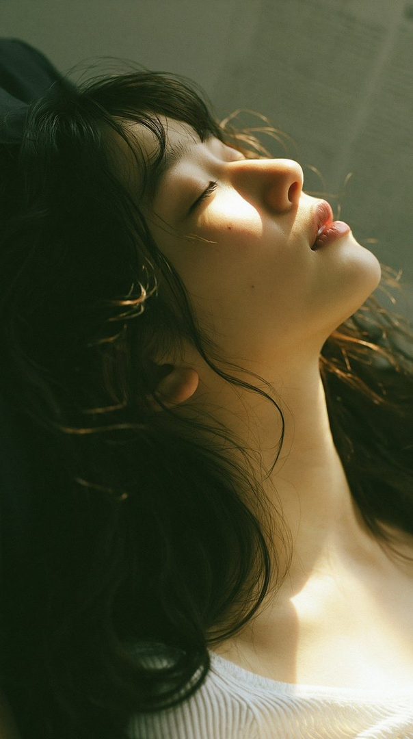 A close-up portrait of a woman with long dark hair, her face tilted upwards with eyes closed, bathed in warm sunlight.