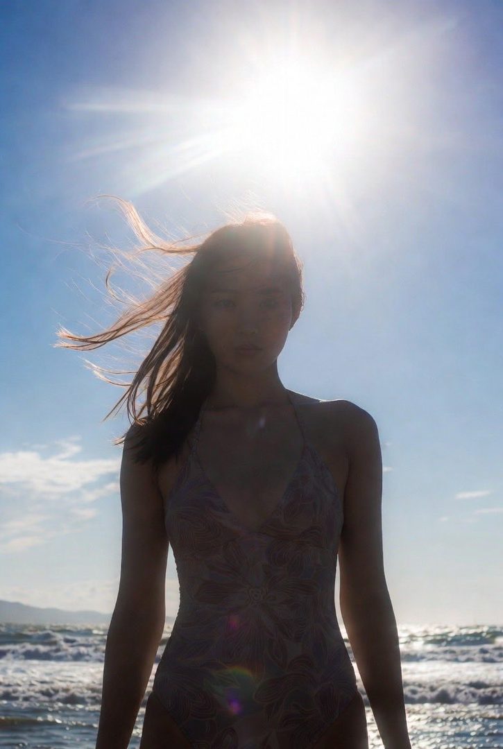 A photorealistic AI-generated image of a woman in a swimsuit standing on a beach with the sun shining brightly behind her.