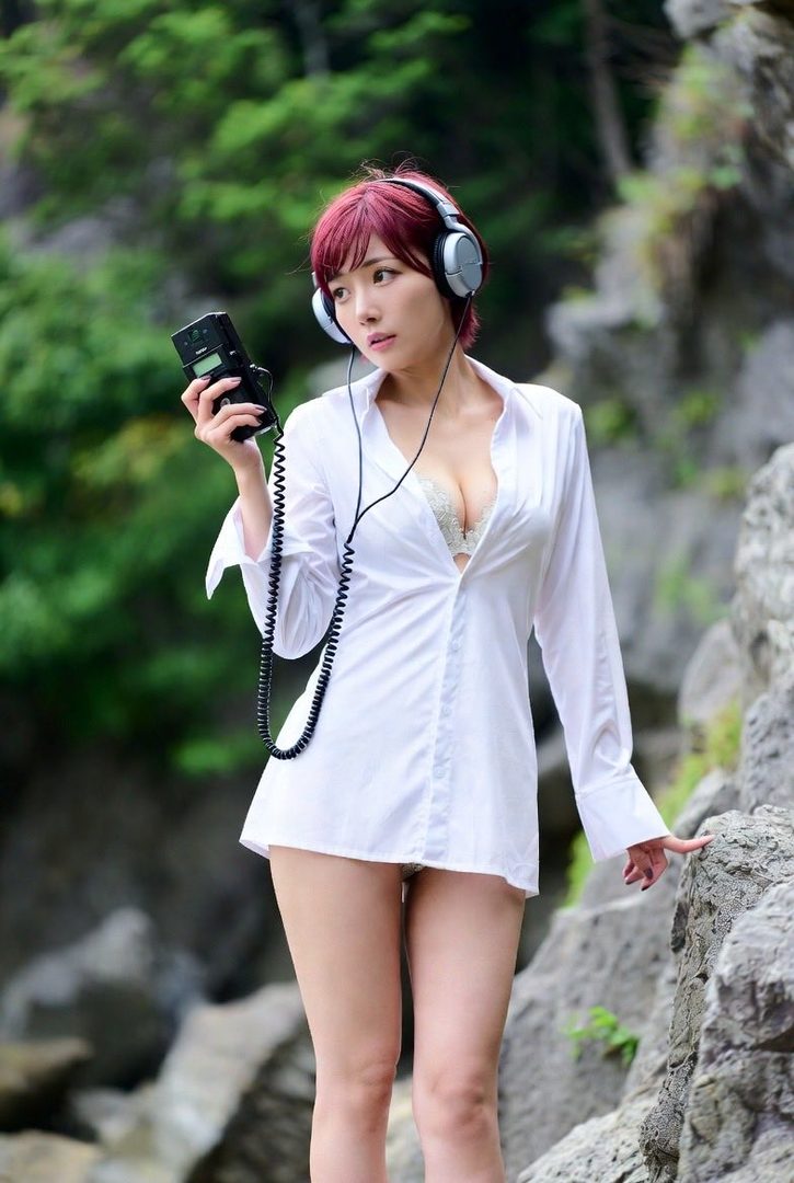 A woman with red hair and headphones wears an unbuttoned white shirt and holds a device outdoors.