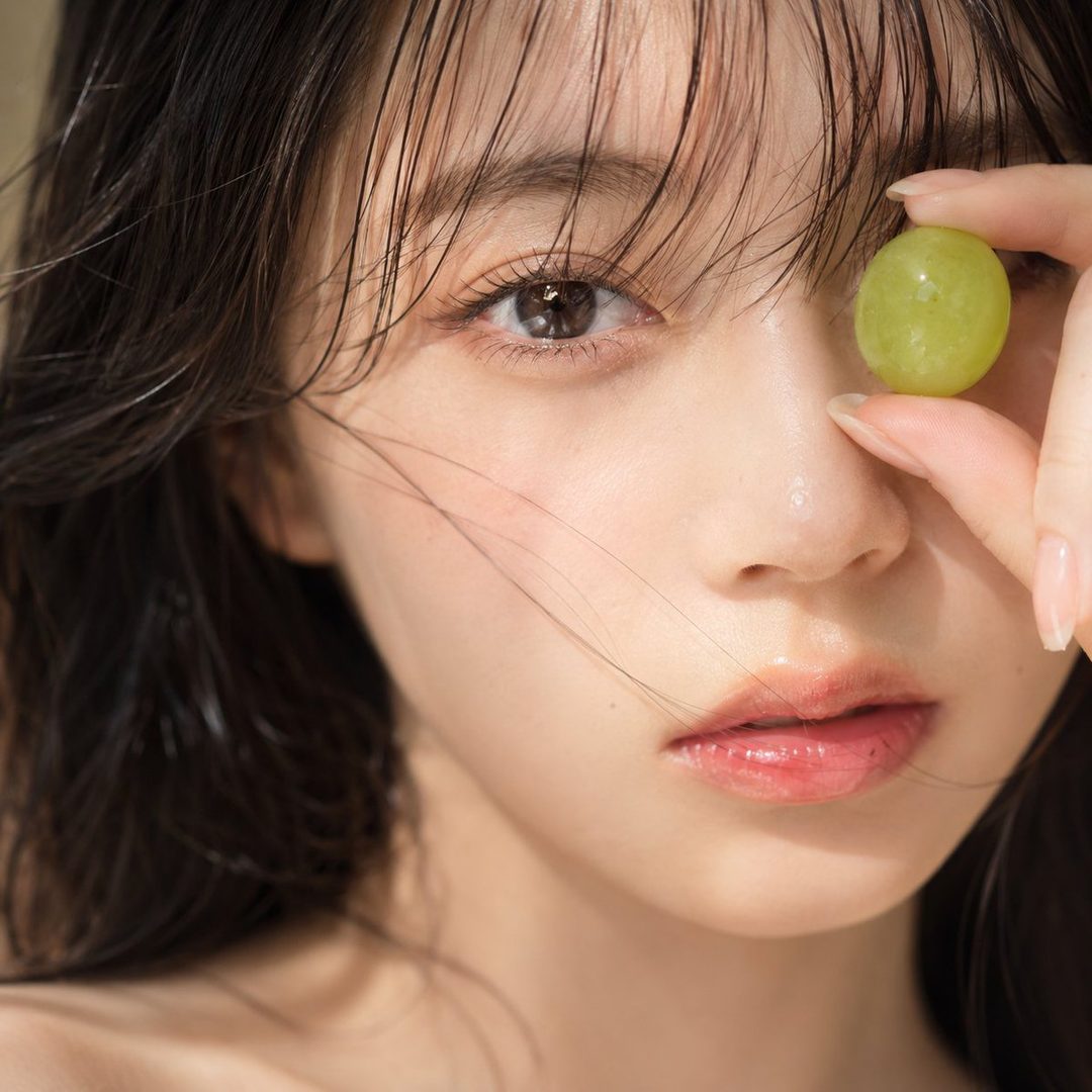 A close-up portrait of a young woman with dark hair and bangs, holding a green grape over one eye.