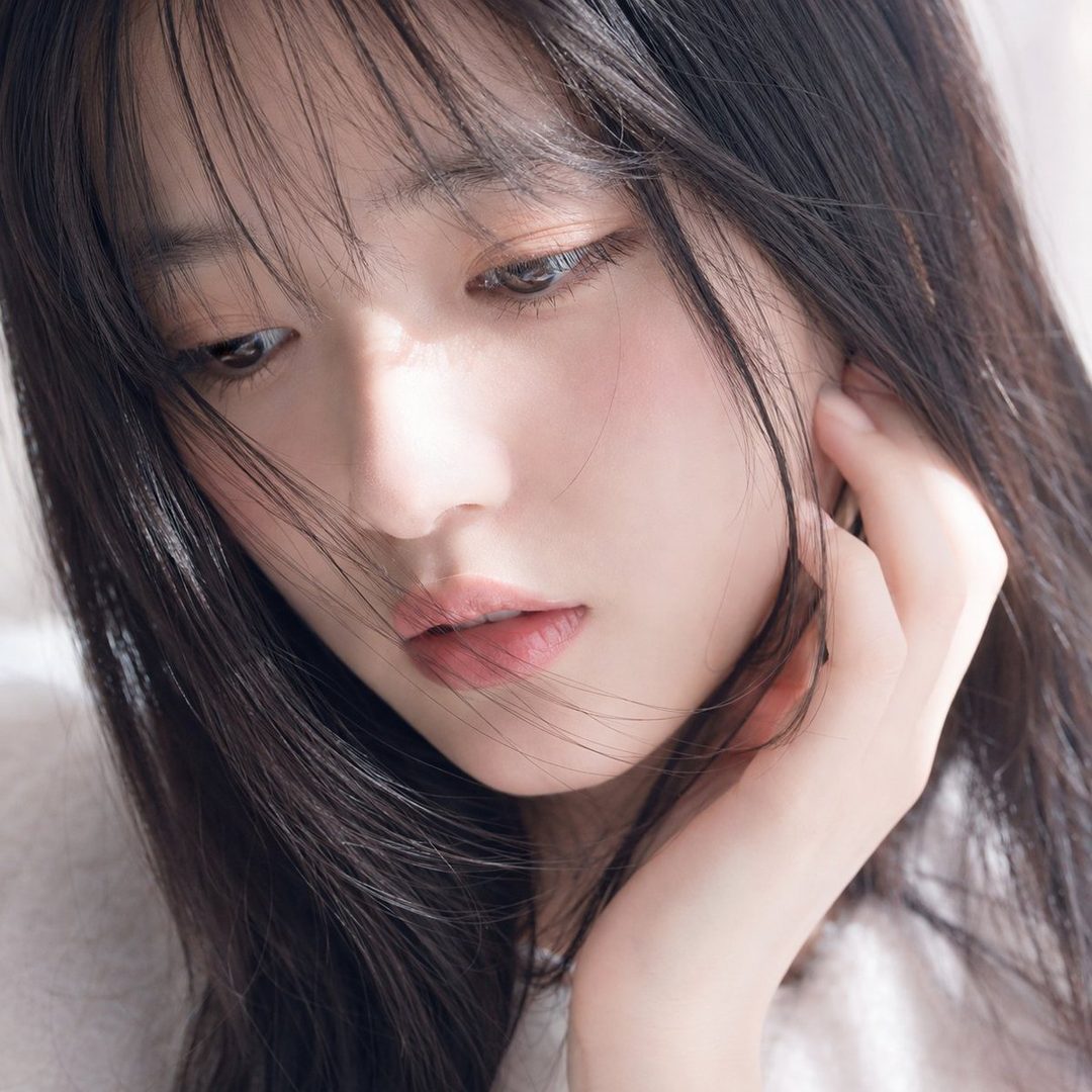 A close-up portrait of a young woman with long black hair and bangs, looking down with a serene expression.