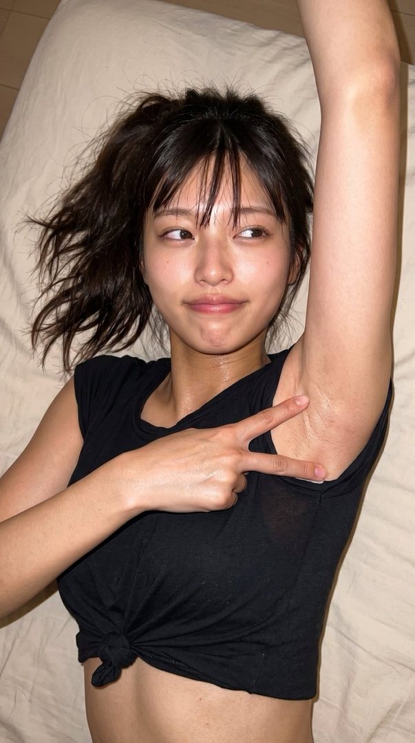 A young woman with brown hair and bangs lies on a bed, pointing at her armpit with a peace sign gesture.