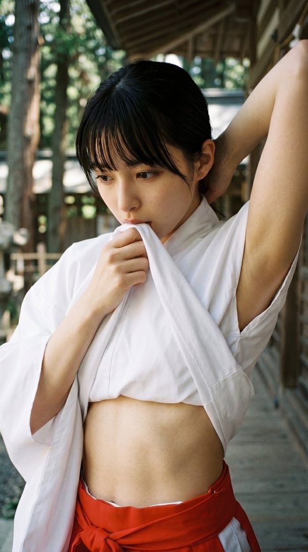 A young woman in a white kimono top and red hakama skirt poses outdoors, looking down with one arm raised.