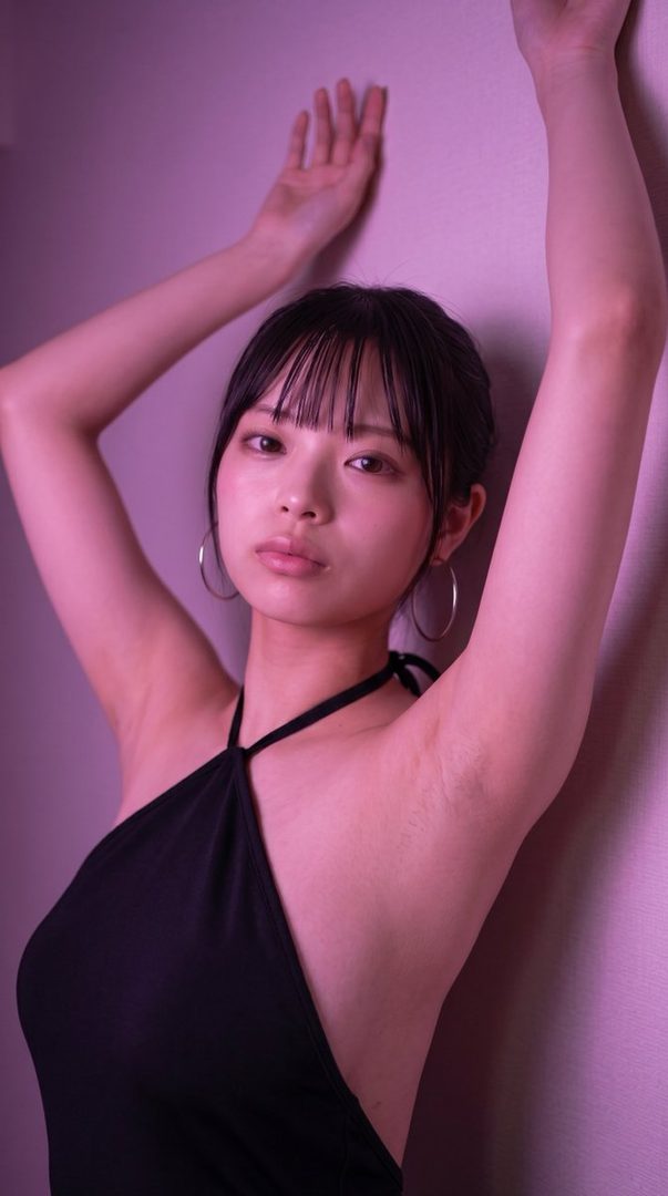 A young woman with black hair and bangs poses against a pink wall, wearing a black halter top and hoop earrings.