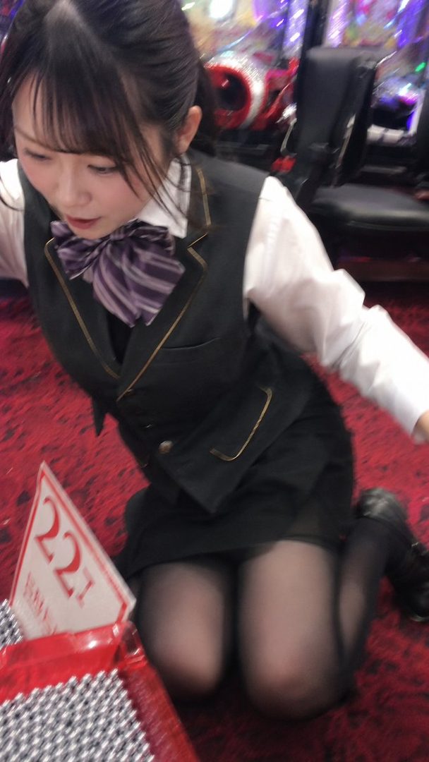 A young woman in a black vest and white shirt with a purple bow tie kneels on a red carpet, looking down at a container of small silver balls.