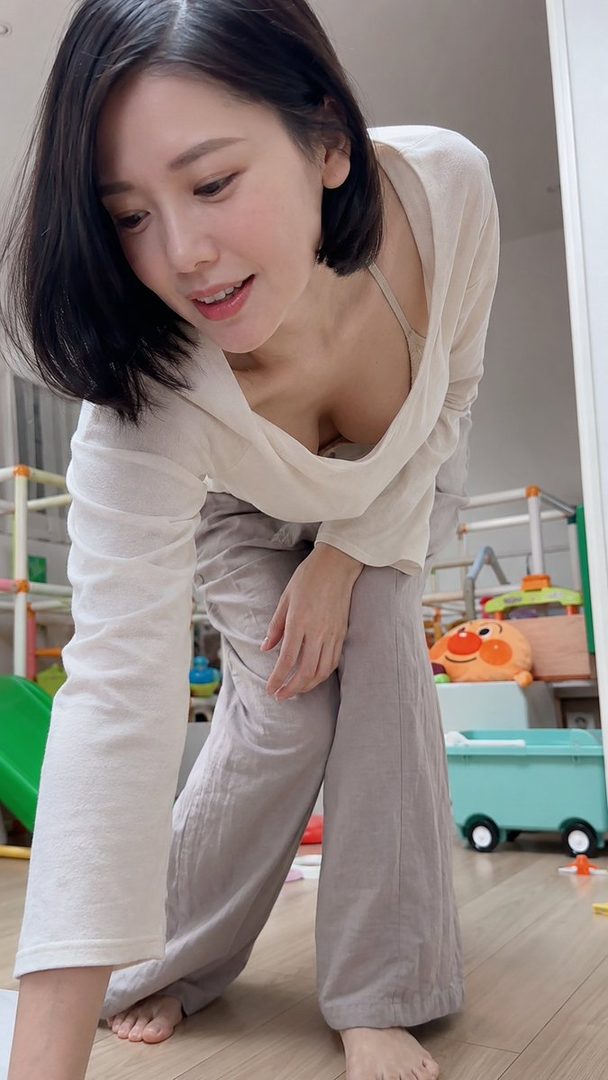A photorealistic image of a Japanese woman with short black hair, wearing a loose white top and grey pants, bending over indoors, showing cleavage.