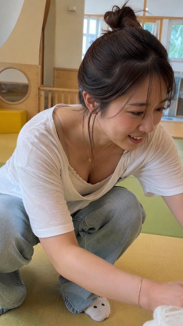 A smiling woman with her hair in a bun, wearing a white t-shirt and jeans, crouches indoors.