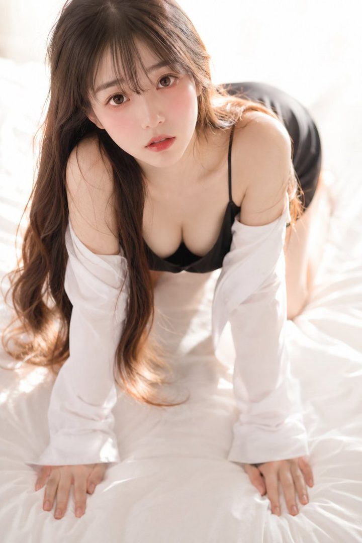 A young East Asian woman with long brown hair and bangs, wearing a black slip dress and an oversized white shirt, kneels on a white bed and looks up at the camera.