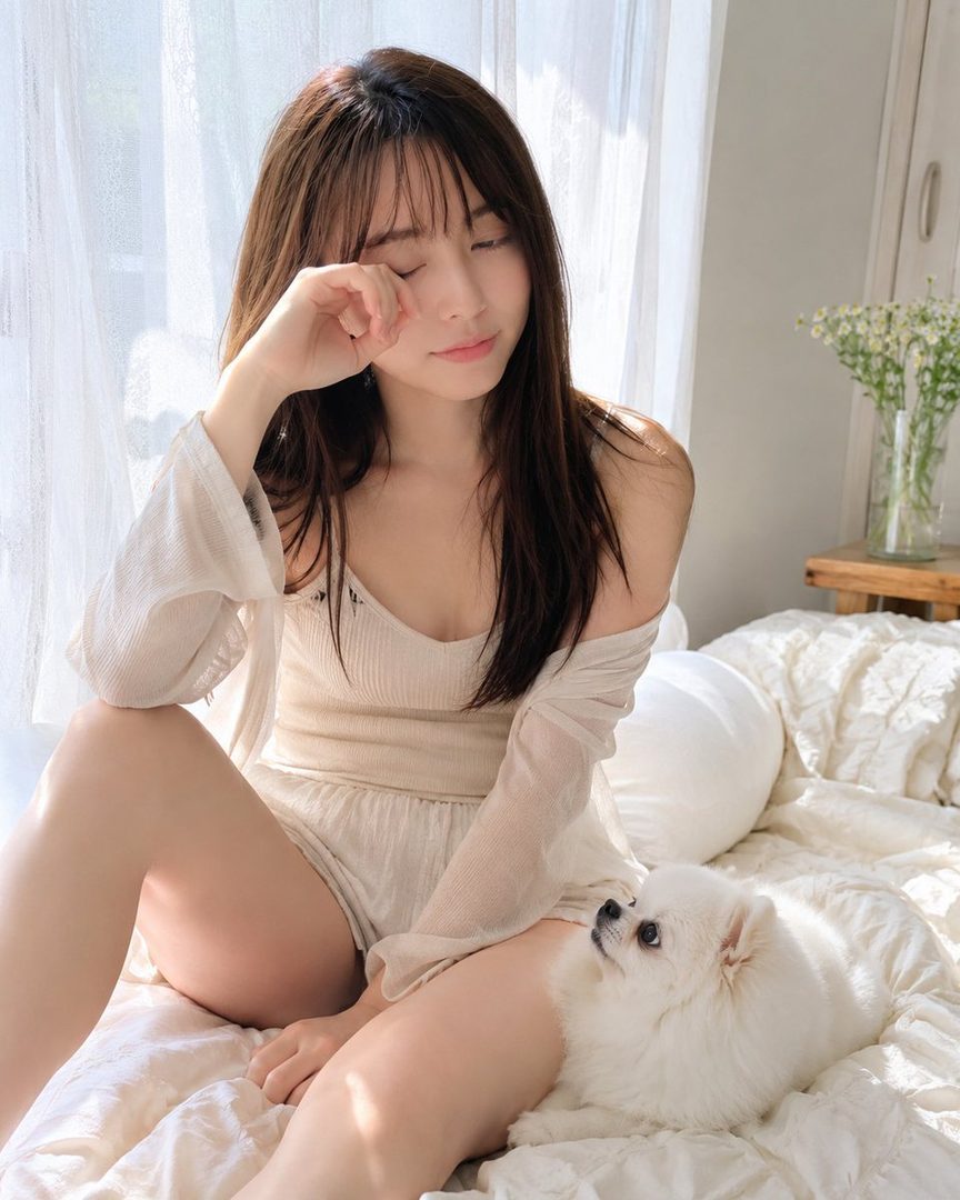 A young woman with long brown hair and bangs, wearing light casual clothing, sits on a bed with a small white dog looking up at her, bathed in soft daylight.