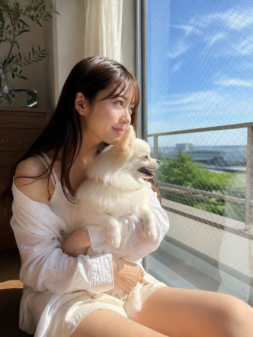 A young woman with long brown hair and bangs, wearing a white shirt and shorts, sits by a window holding a small white dog, looking out at the sunny sky.