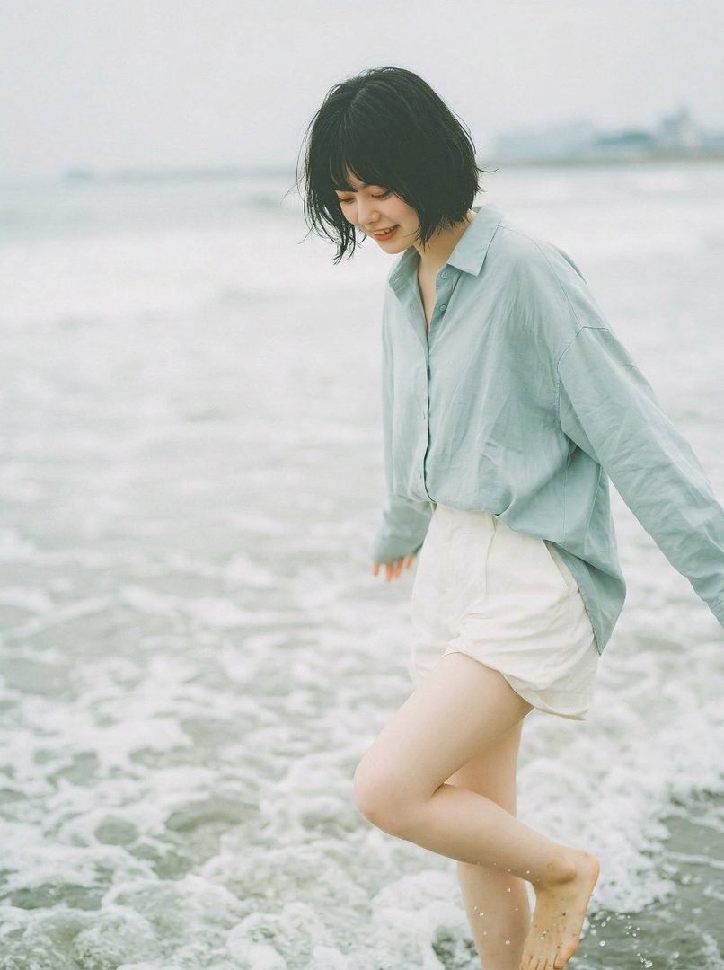 A young woman with short black hair and bangs, wearing a light blue shirt and white shorts, walks barefoot in the shallow water at the beach, smiling softly.