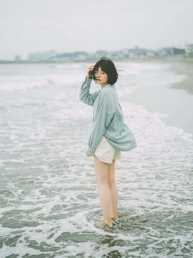 A young woman with short black hair stands in the shallow water at a beach, looking back at the camera.