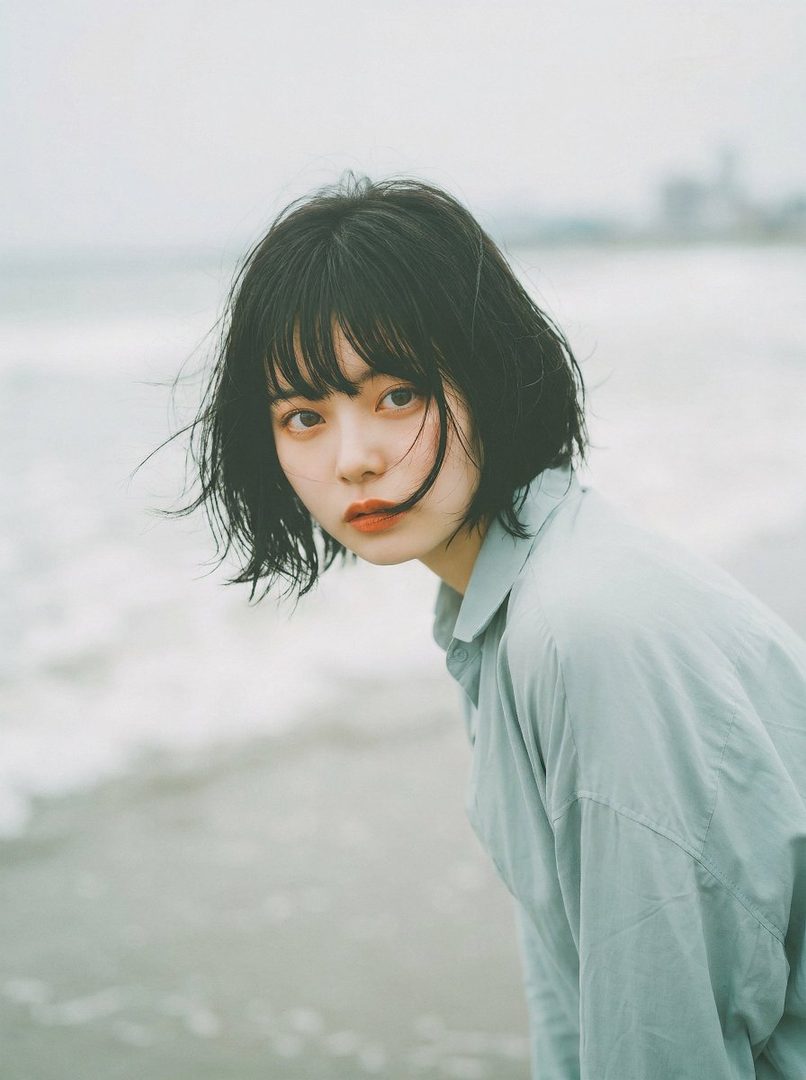 A young woman with short black hair and bangs looks directly at the camera while standing by the ocean.