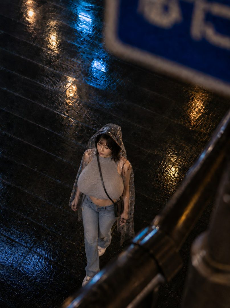 A woman in a clear raincoat and grey crop top walks on a wet street at night, with reflections of lights on the ground.