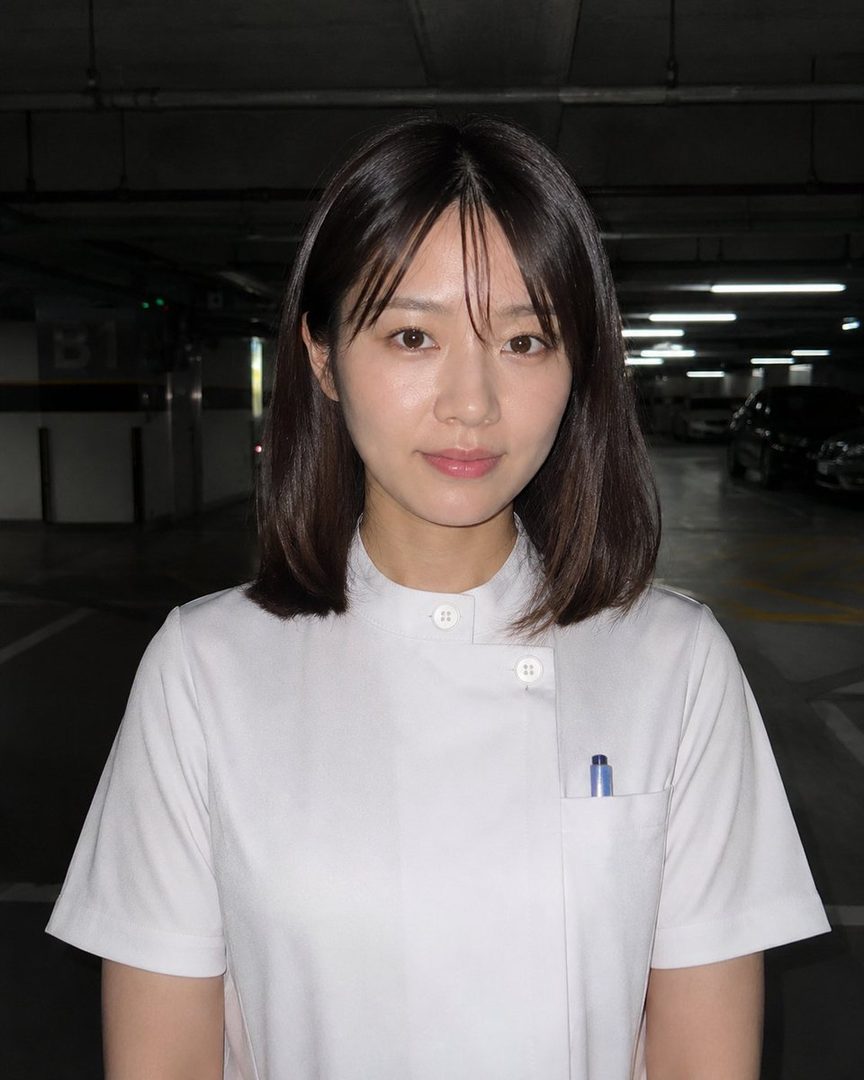 A woman with short brown hair and bangs, wearing a white uniform, stands in an indoor parking garage.