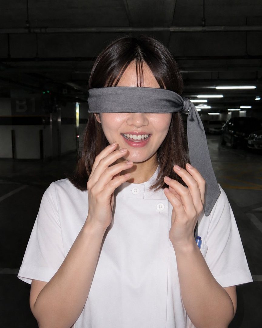 A young woman with a blindfold over her eyes smiles and holds her hands near her face in an indoor parking garage.