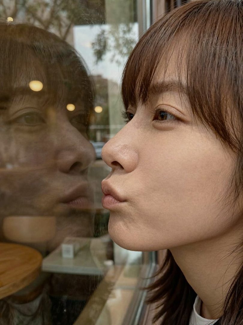 A woman with brown hair and bangs makes a kissing face at her reflection in a window.