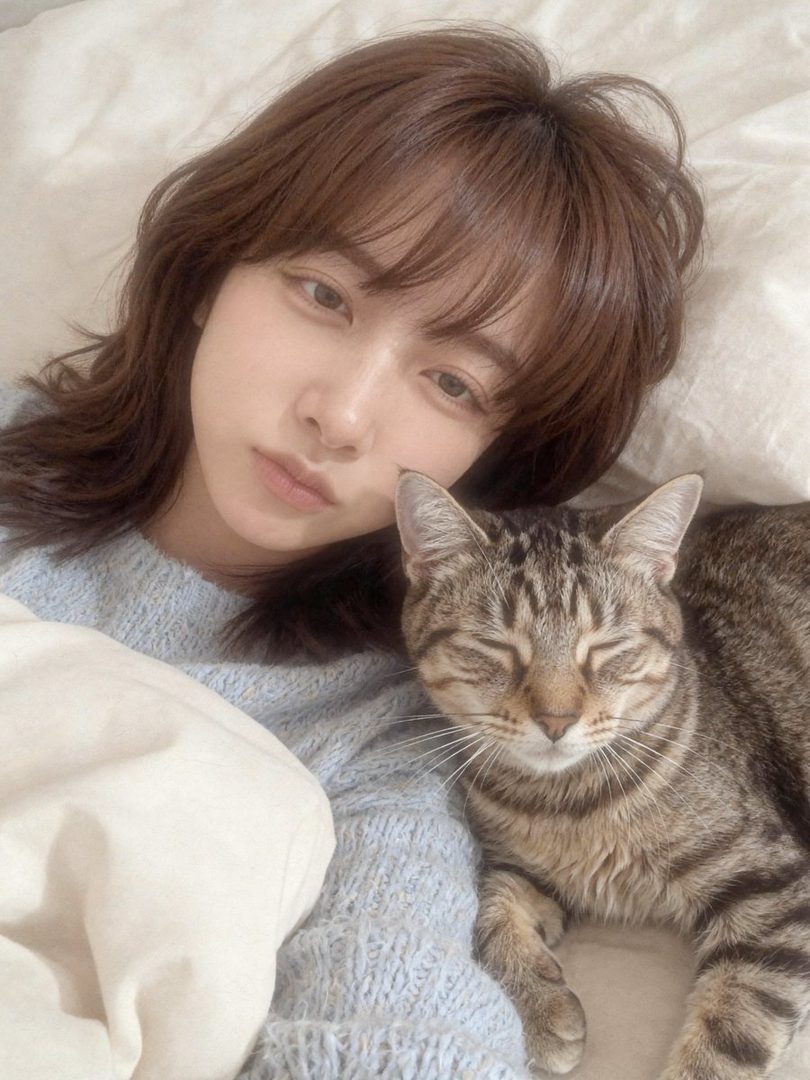 A young woman with brown hair and bangs lies in bed next to a sleeping tabby cat, wearing a light blue knit sweater.