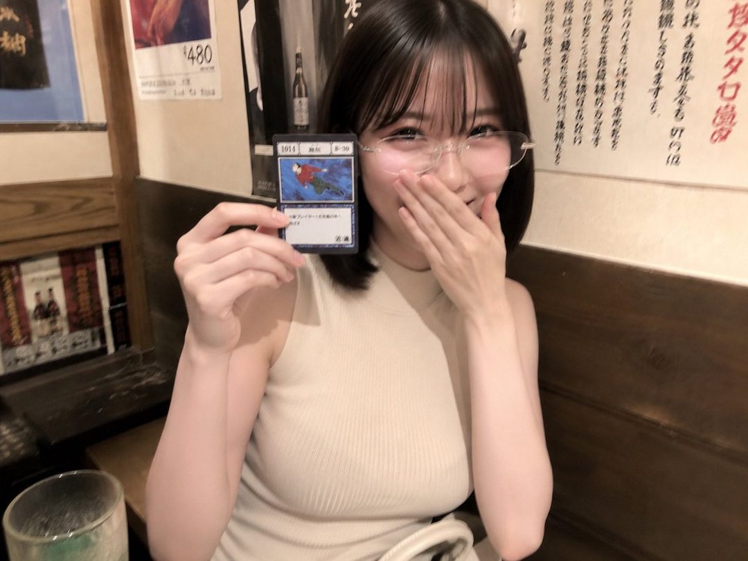 A young woman with short black hair and glasses smiles while holding a trading card, covering her mouth with her hand in an indoor setting.