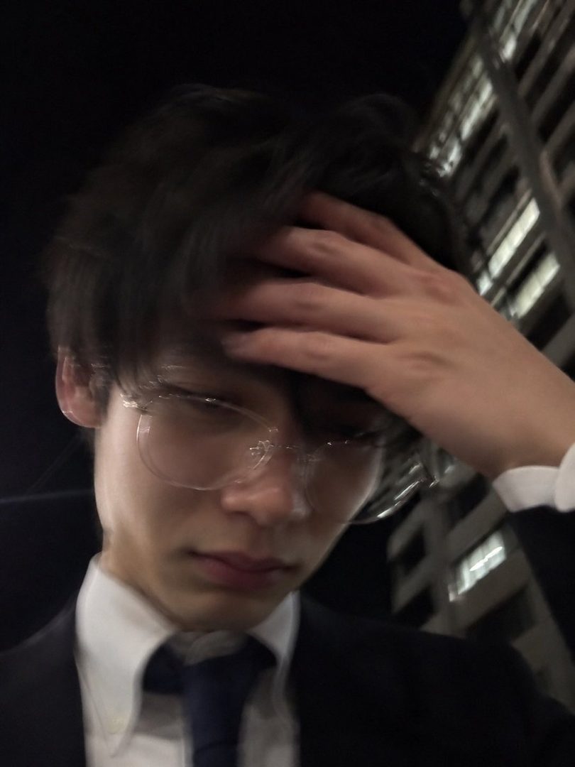 A young man in a suit and glasses, with his hand on his head, stands in a blurry city at night.