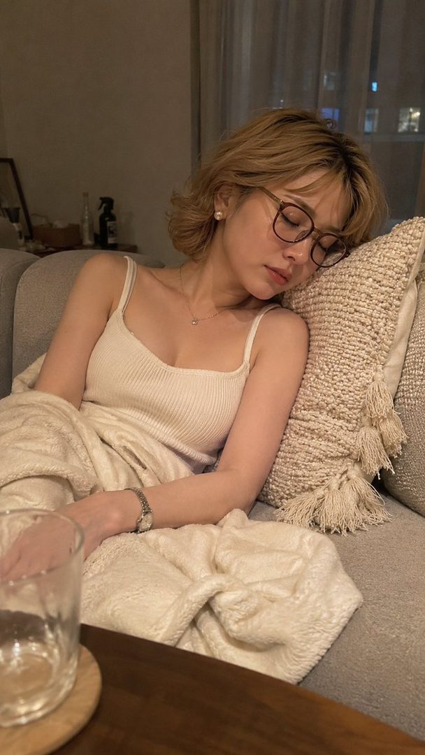 A photorealistic AI-generated image of a woman with short blonde hair and glasses, sleeping peacefully on a sofa indoors at night.