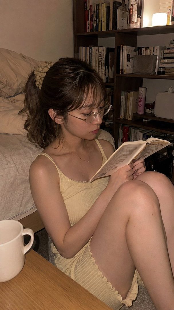 A young woman with glasses and brown hair in a ponytail is sitting on the floor, reading a book in a cozy indoor setting at night.