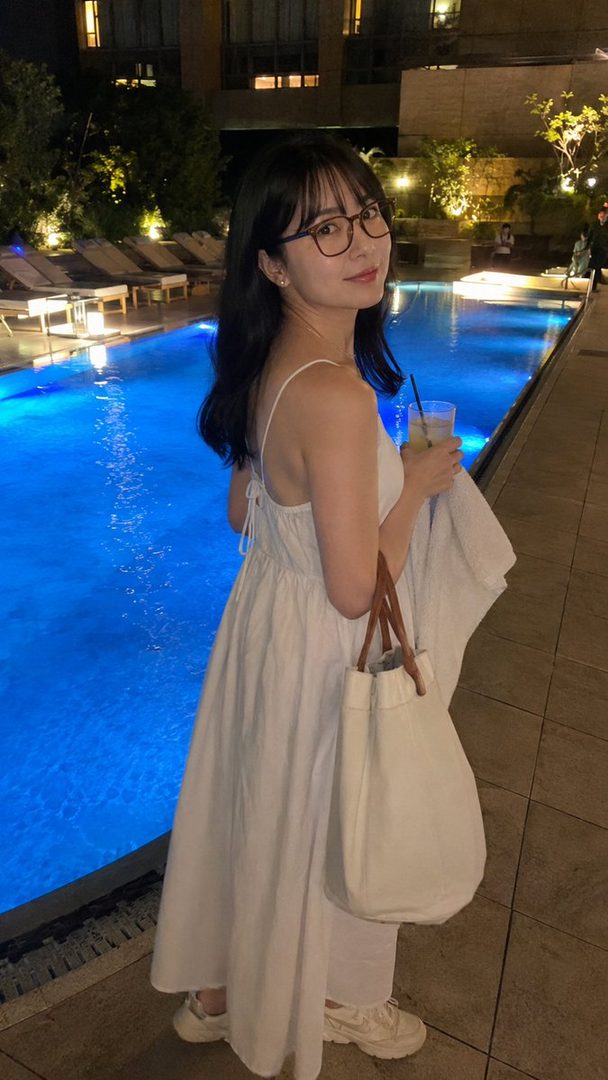 A young woman in a white dress and glasses stands by a swimming pool at night, holding a drink and a white bag.