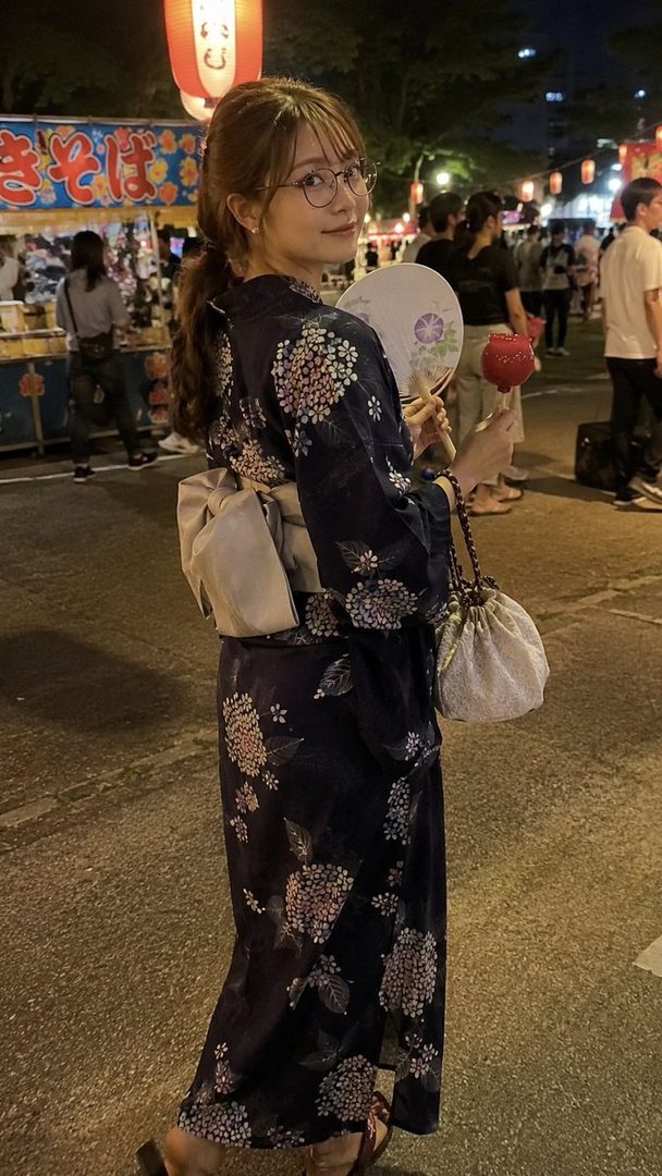 A young woman in a dark blue yukata with floral patterns, holding a fan and a candy apple, looks back over her shoulder at a night festival.