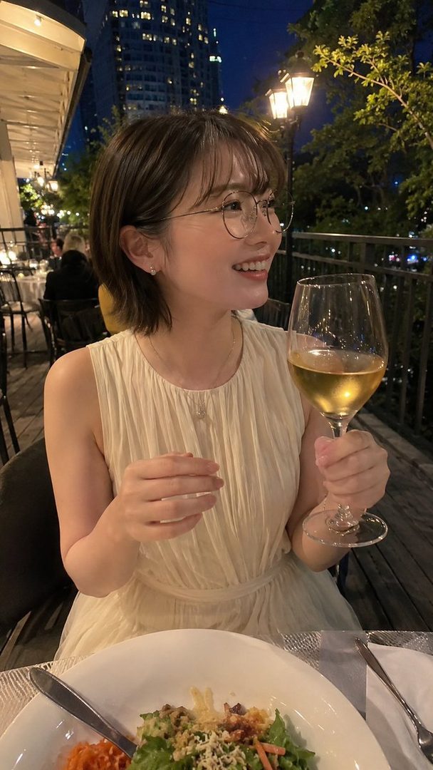 A smiling woman with short brown hair and glasses holds a glass of white wine at an outdoor restaurant with city lights in the background at night.