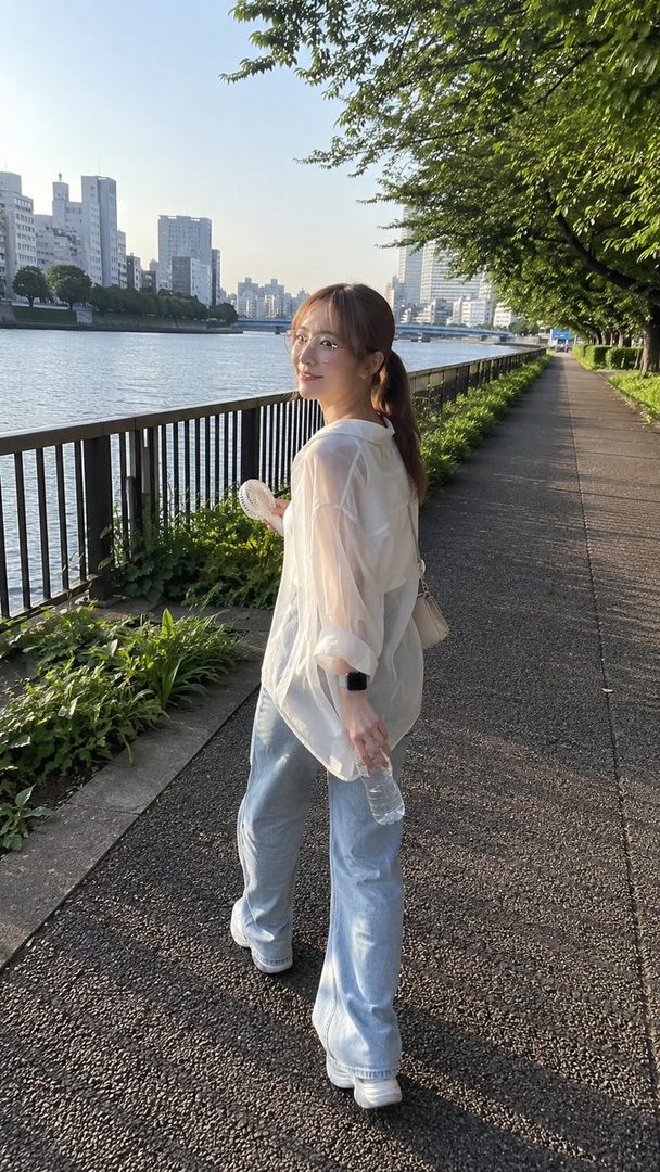 A young woman with a ponytail and glasses walks along a river path in a city during golden hour, looking back over her shoulder.