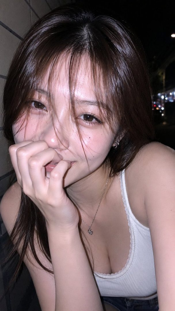 A young woman with long brown hair and bangs smiles playfully at the camera, biting her finger, wearing a white tank top at night.