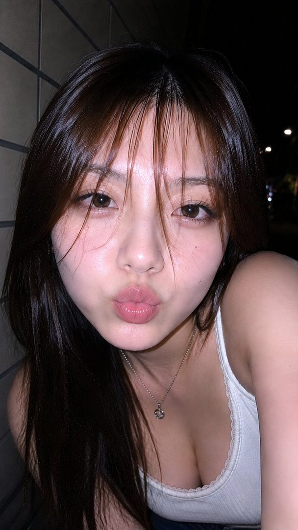 A young woman with long brown hair and bangs, wearing a white knit top, pouts at the camera in a close-up outdoor shot at night.