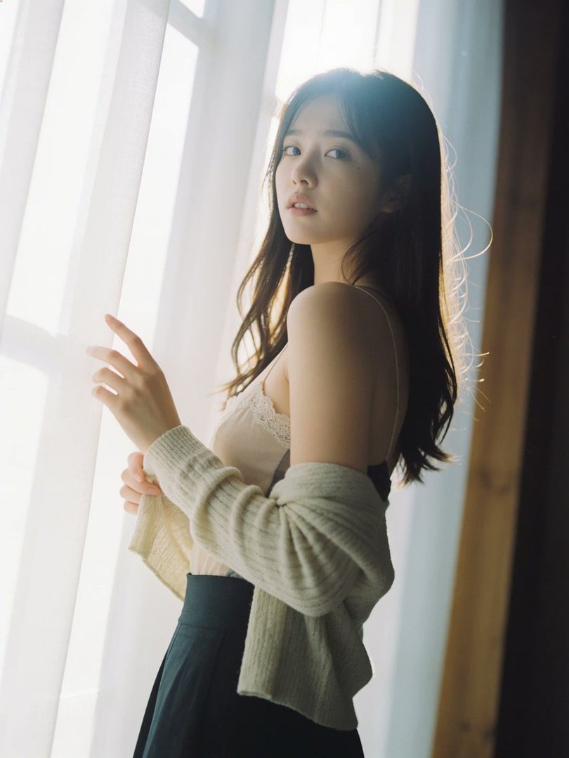 A young woman with long dark hair stands by a window, looking over her shoulder with soft light filtering through the curtains.