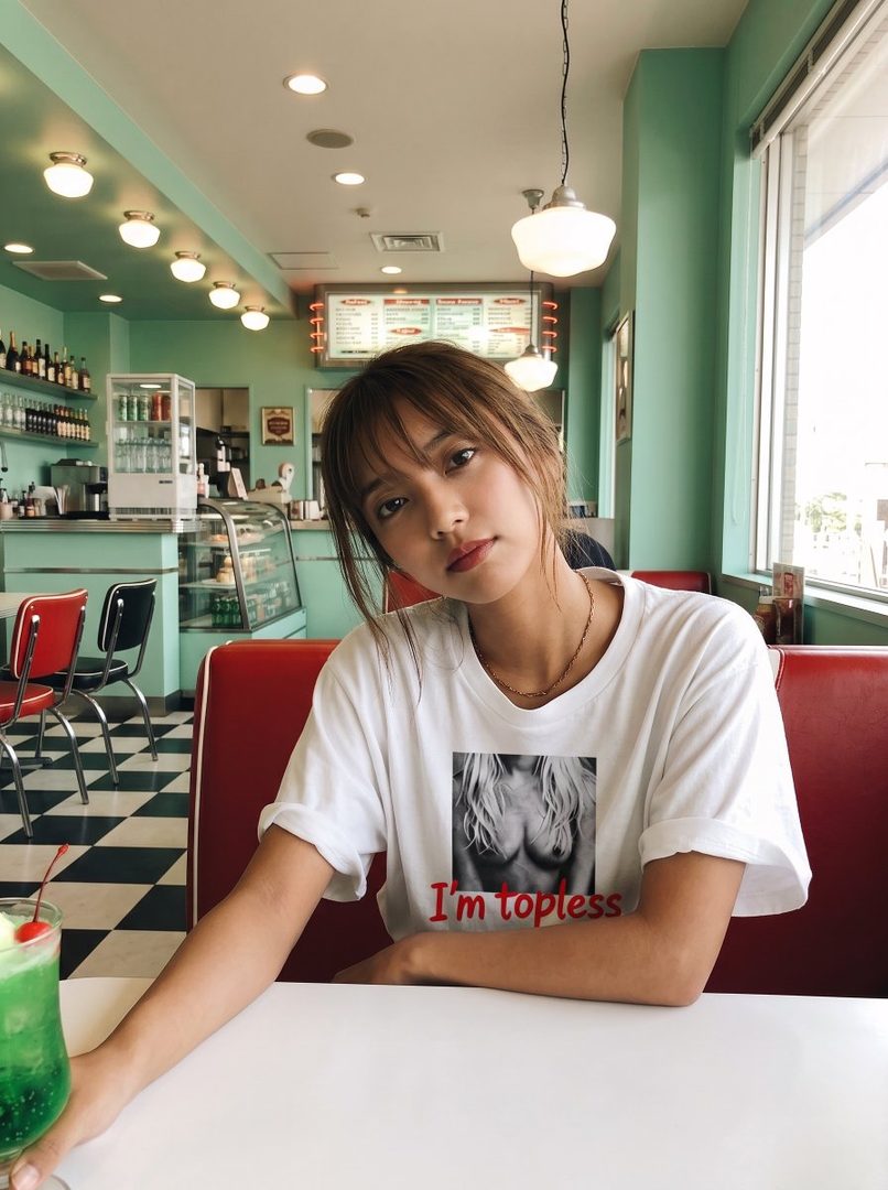 A young woman with brown hair and bangs sits at a diner booth, wearing a white t-shirt with a suggestive graphic.